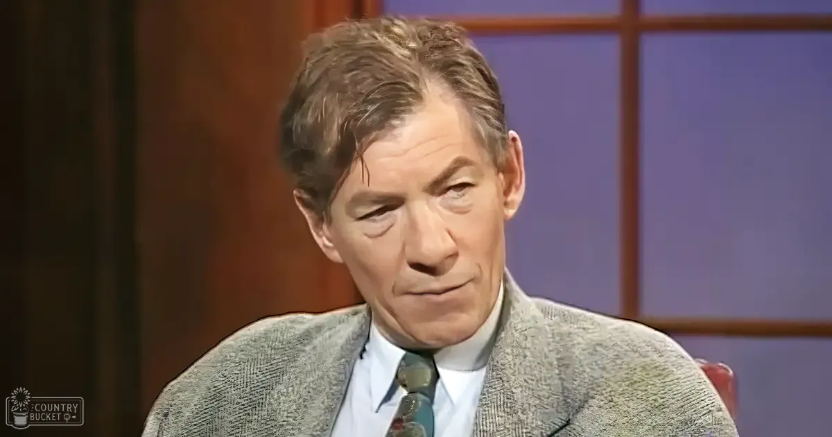 An older adult male wearing a formal blazer and dress shirt with tie, seated against a backdrop of wooden paneling and a window-like design in a studio setting.