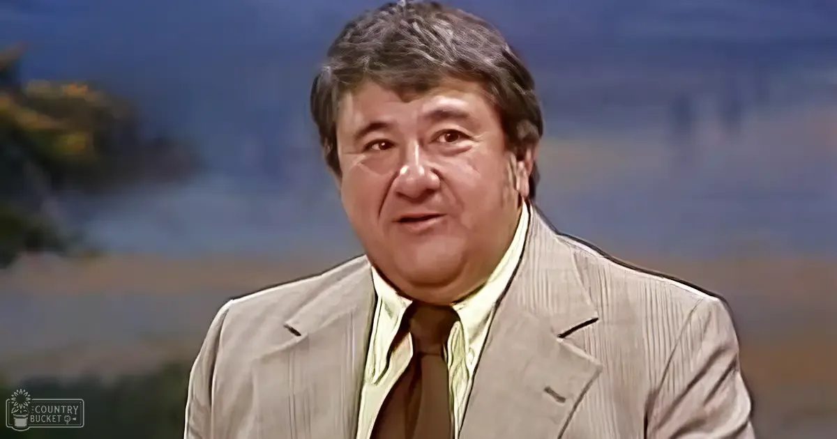 An older adult male wearing a light-colored striped suit jacket and collared shirt against a blurred natural backdrop in what appears to be a studio setting, suggesting an interview or talk show environment.