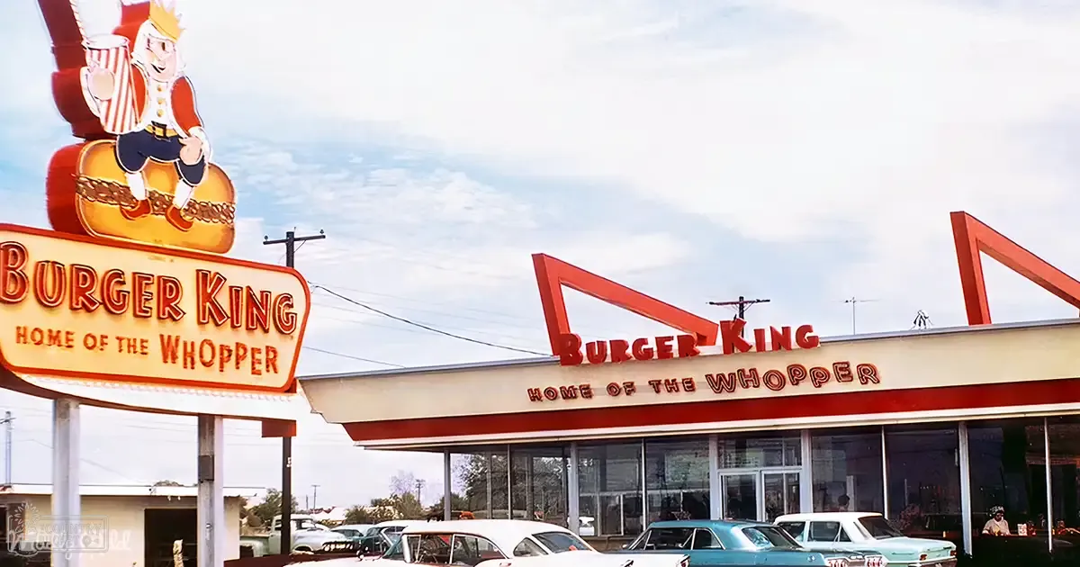 Vintage cars parked outside a Burger King restaurant with large glass windows, red sign, and illuminated animated character atop burger. Urban area, partly cloudy sky, clear day.