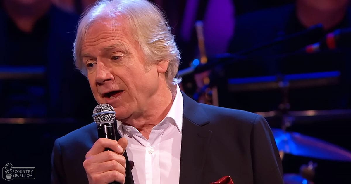 An older adult male, possibly in his 60s or 70s, wearing a dark suit jacket over a white button-up shirt with a classic collar and crisp cotton material, holding a microphone on stage at Madly Odd concert setting under artificial stage lighting.