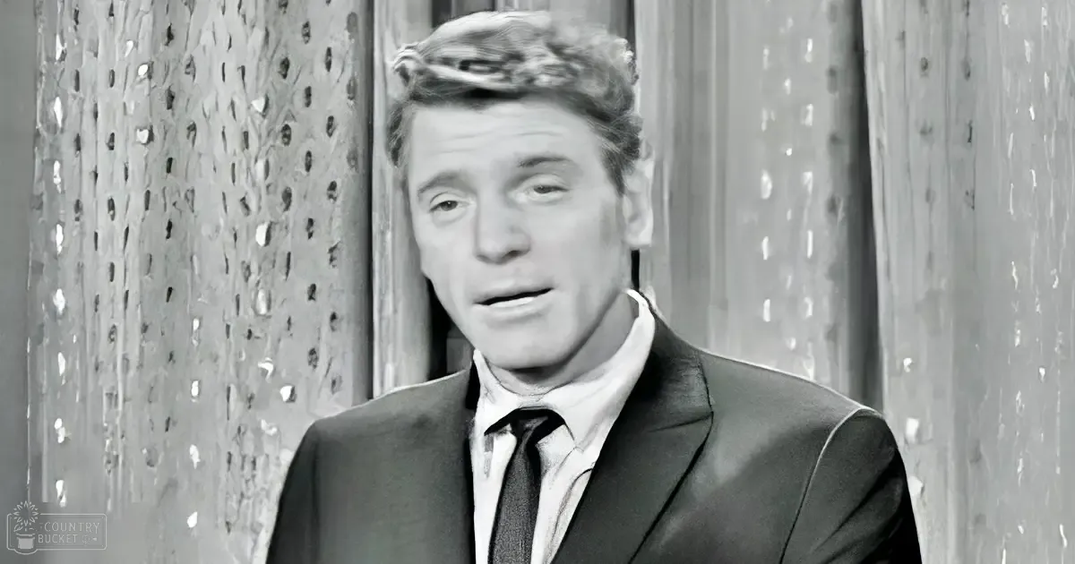 Middle-aged adult male in a dark suit jacket, white dress shirt, and dark tie against a textured wall backdrop.