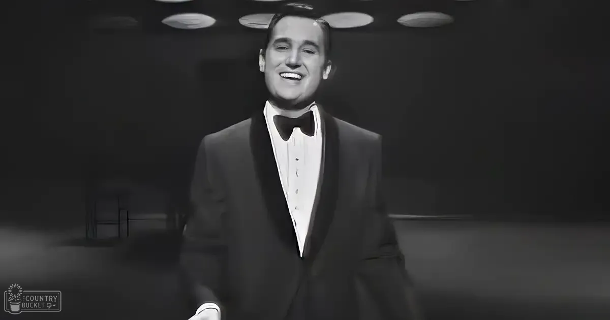A man in an adult age band wearing a formal tuxedo with a classic cut and white shoes is seen indoors under dimly lit ceiling lights.