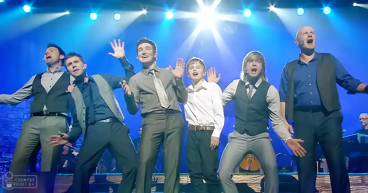 Group singing performance at an indoor concert or theater setting with children and young adults wearing formal attire under bright stage lighting.