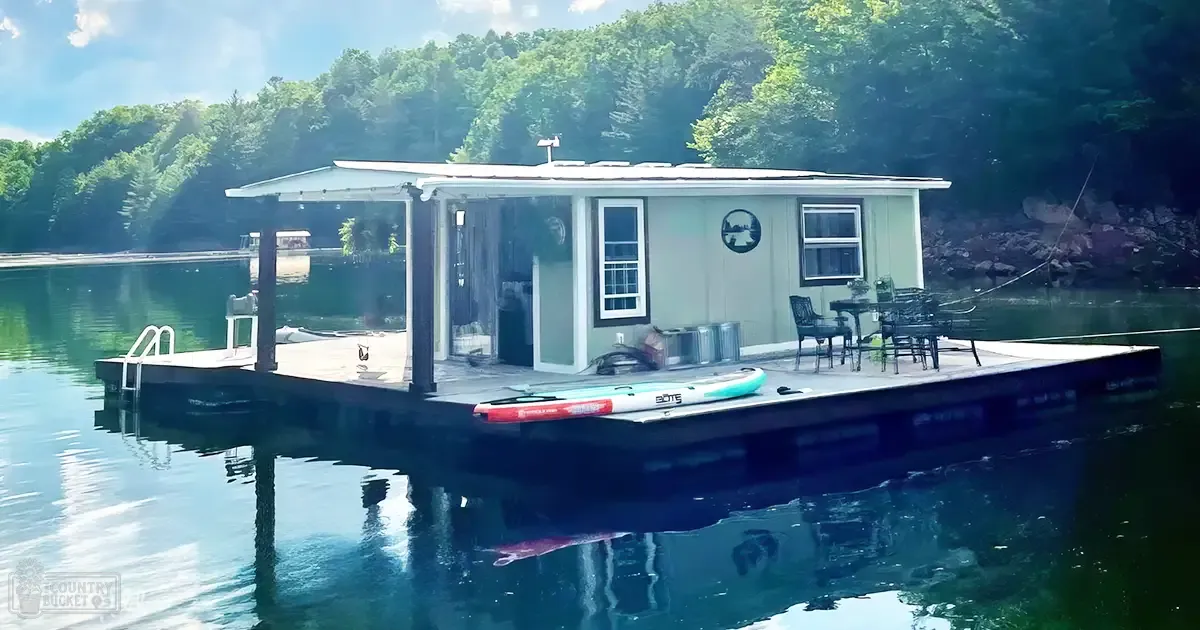 A peaceful lakeside scene with a floating house, outdoor furniture, and paddleboards on the dock under lush greenery.