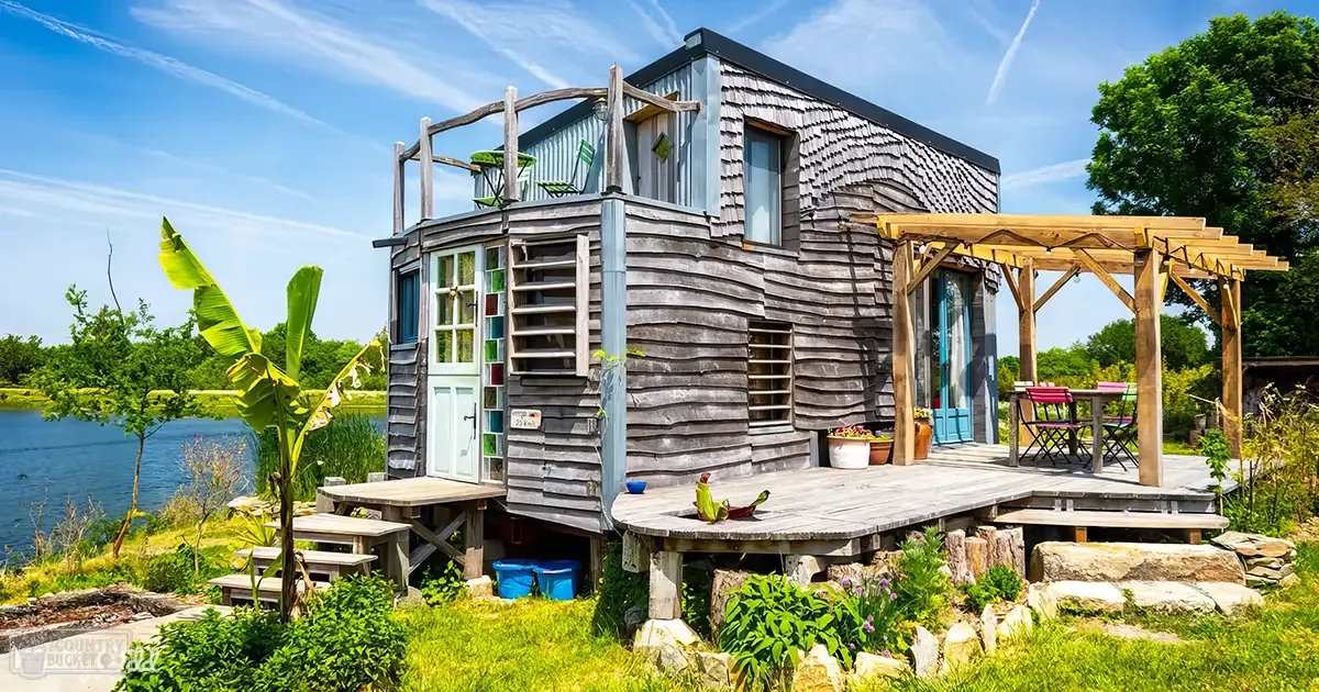 A rustic wooden cabin surrounded by lush greenery near a serene body of water, with outdoor furniture under a pergola and potted plants adding color to the scene.