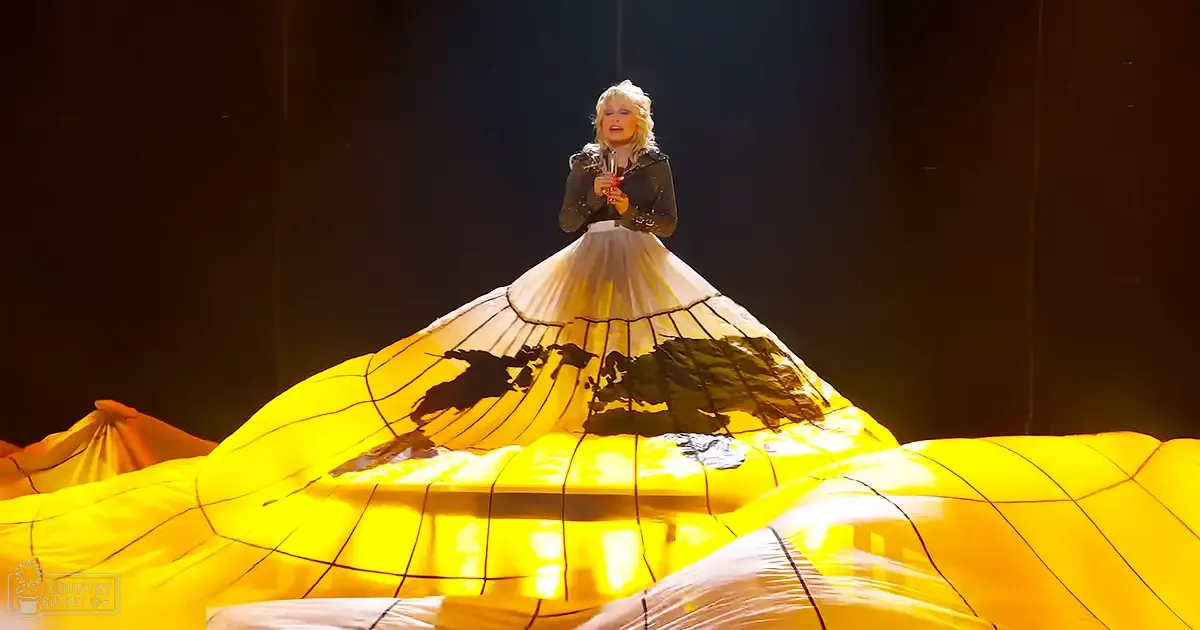 A middle-aged female performer in a concert hall, wearing an artistic white skirt with black trim and intricate animal silhouettes, standing beneath a large yellow fabric structure under dim stage lighting.