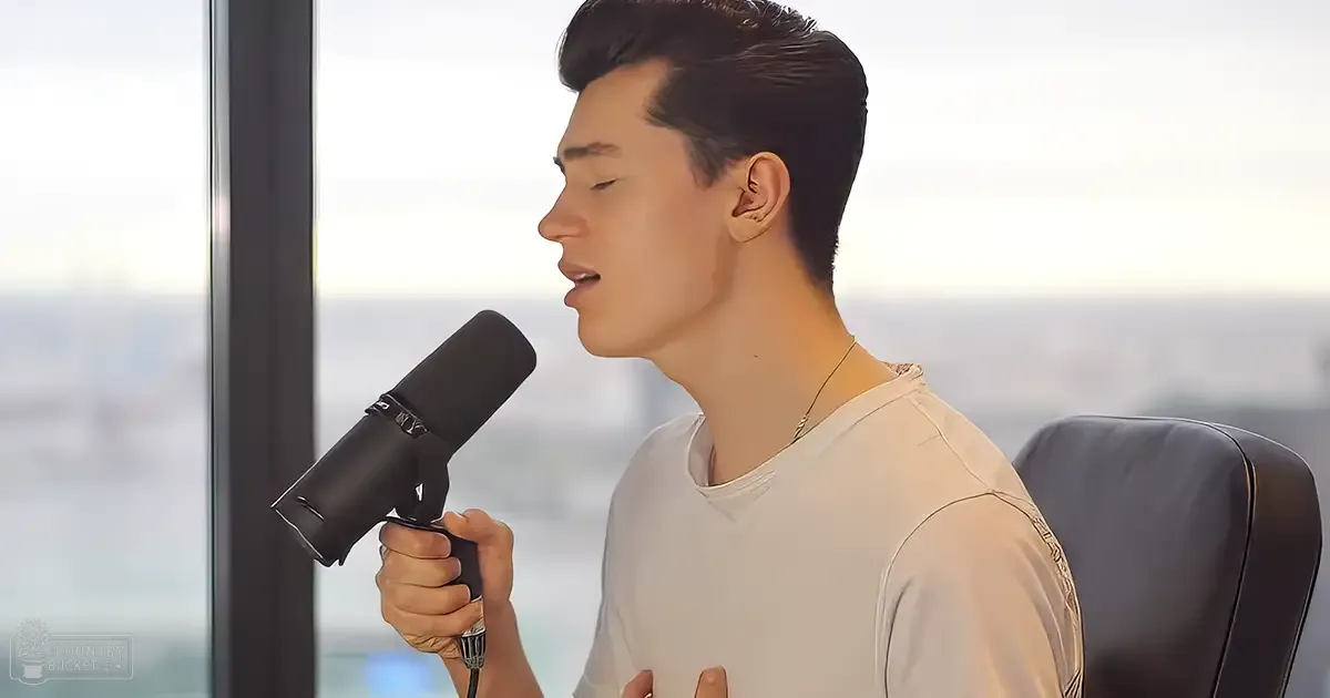 A young adult male singer in a relaxed fit white long-sleeved shirt, wearing a thin gold chain around his neck, singing into a professional microphone with large windows showing an outdoor view suggesting daytime and possibly spring or summer.
