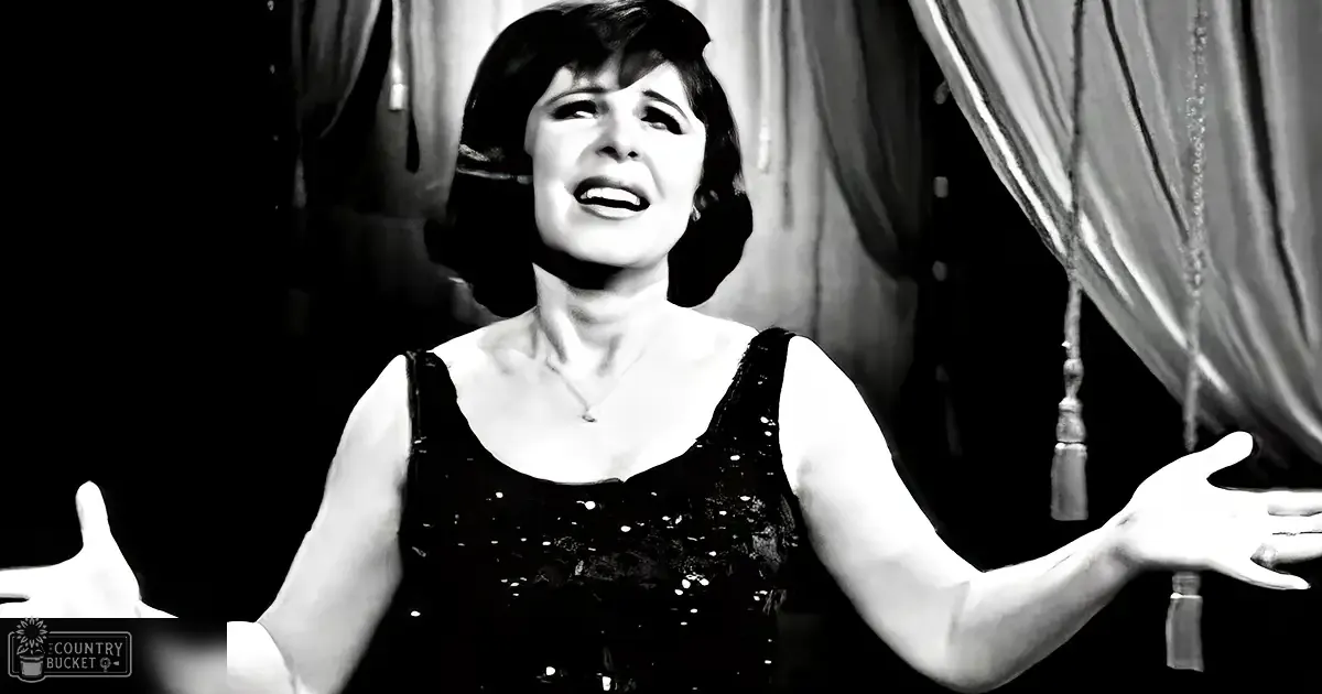 A middle-aged adult female wears a black, sequined sleeveless top on stage or performance area with curtains and tassels.