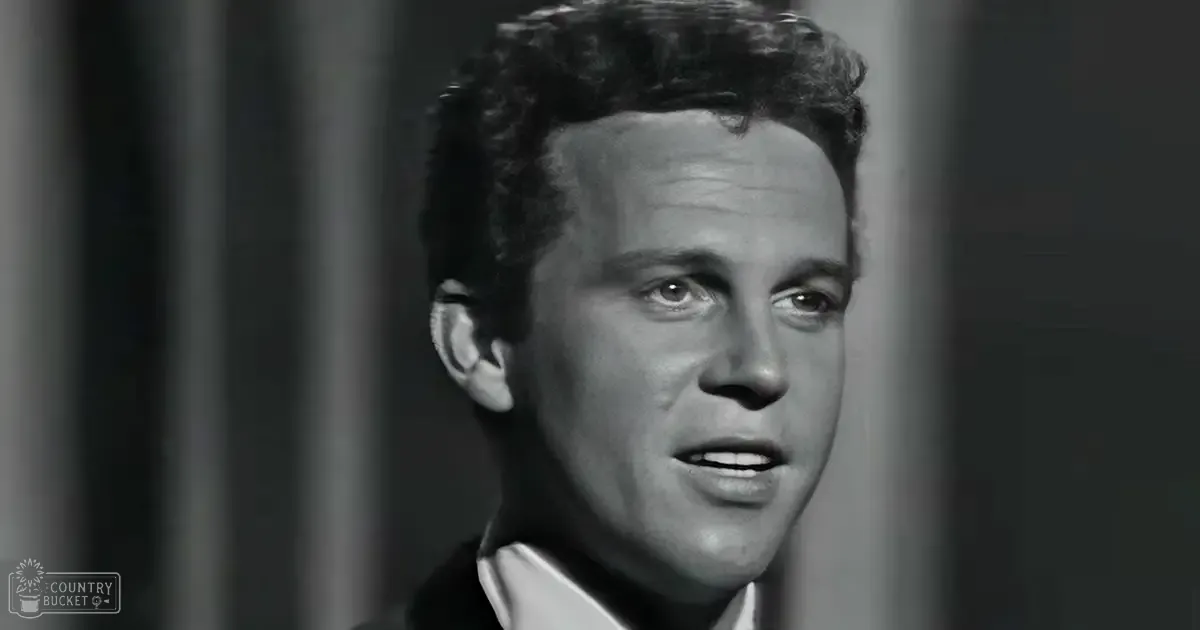 Young adult male wearing a dark suit jacket with crisp white shirt against neutral-colored curtains in a studio setting.