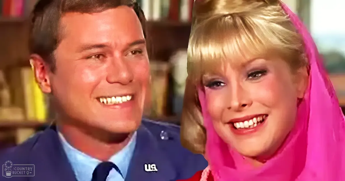 A young adult male in a dark blue military-style jacket with 'U.S.' patch, light blue shirt, and dark tie stands beside an adult female wearing a bright pink dress or costume. The setting appears to be an indoor library or study with bookshelves visible.