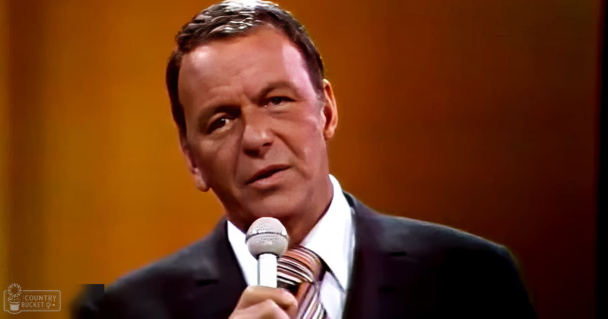 A middle-aged adult male wearing a dark suit jacket with a striped tie in an indoor setting with warm lighting and an orange curtain backdrop.