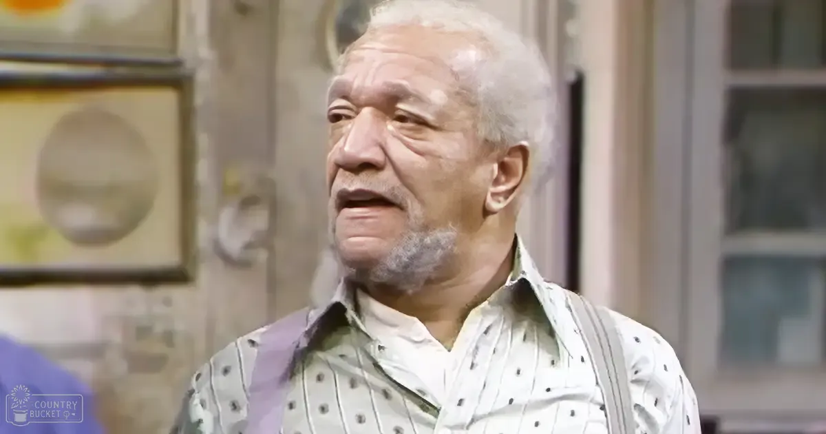 An older adult male wearing a white shirt with vertical stripes and small black dots, paired with light-colored suspenders with darker polka dots, stands indoors in a home-like environment. Natural light streams through the window, suggesting it might be daytime.