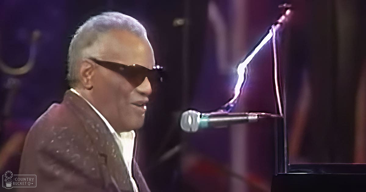 An older adult male playing a piano on an indoor stage with dark curtains and focused lighting, wearing a patterned blazer over a collared shirt and tie or scarf.