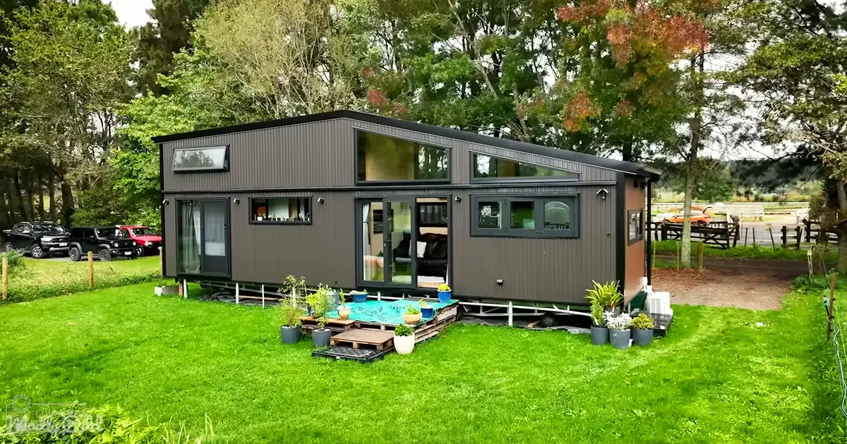 A modern tiny house with dark gray siding and large windows, surrounded by potted plants on wooden pallets and parked vehicles in a rural setting.