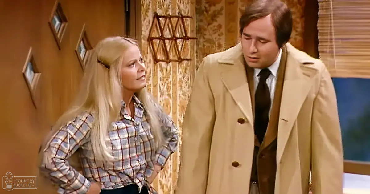 A young adult woman wearing a plaid shirt with red, white, and blue patterns indoors; a middle-aged man in a beige trench coat over a suit stands nearby. The setting is an indoor environment with natural light.
