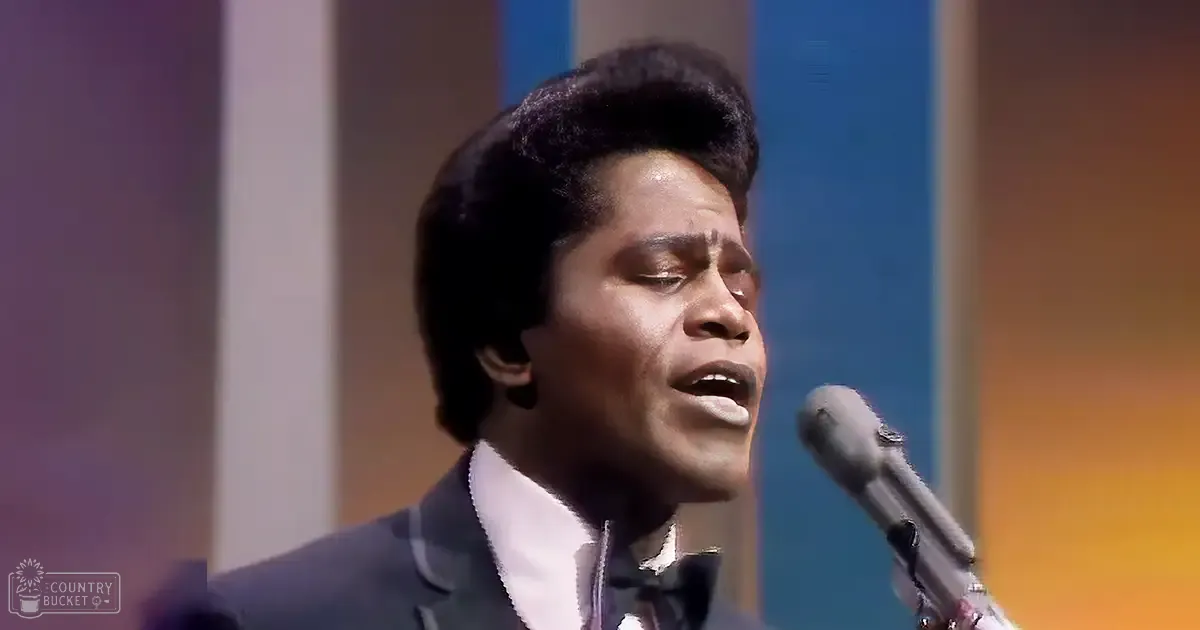 A young adult male in a tailored dark blue suit jacket with a white collared shirt and bow tie, singing into a microphone in a studio setting.