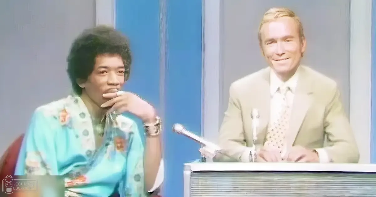 A young adult male in a casual light blue floral-patterned outfit sits next to an older adult male wearing a beige suit jacket, both seated at a table with microphones andcripts, suggesting an interview or talk show setting. The indoor studio has minimalist decor with blue panels as the backdrop.