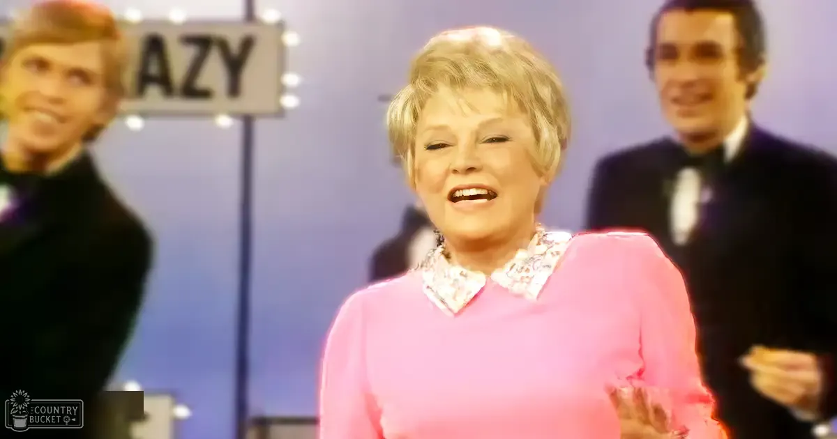 A middle-aged woman in a pink floral-patterned sweater stands indoors, possibly at an event or studio setting with two men wearing dark suits on either side.
