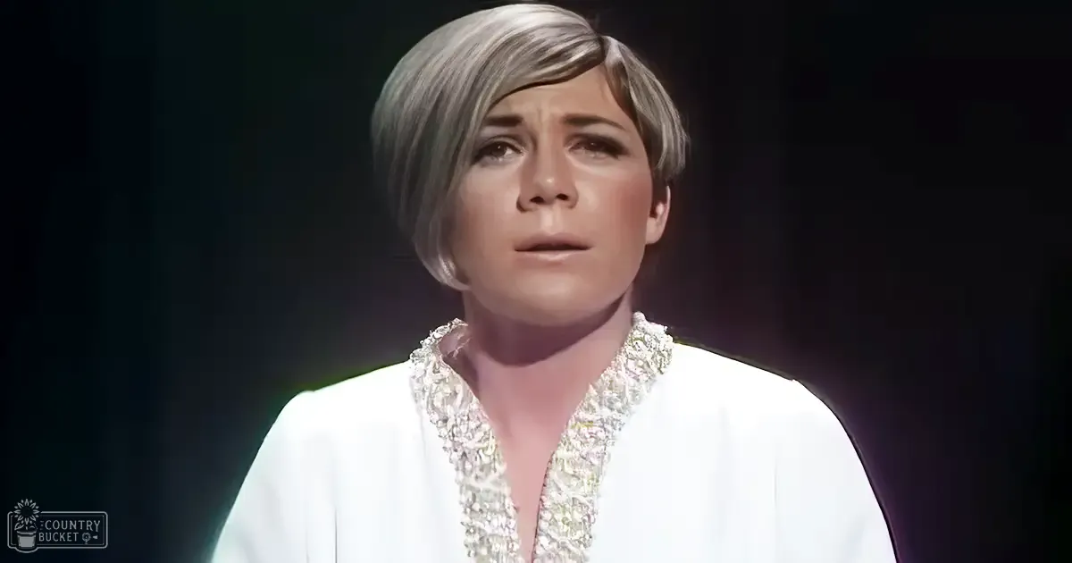 A middle-aged woman in an indoor setting wearing a white garment with intricate embellishments around the neckline and shoulders, V-neckline, fitted cut, short sleeves, light fabric suitable for formal or evening wear.