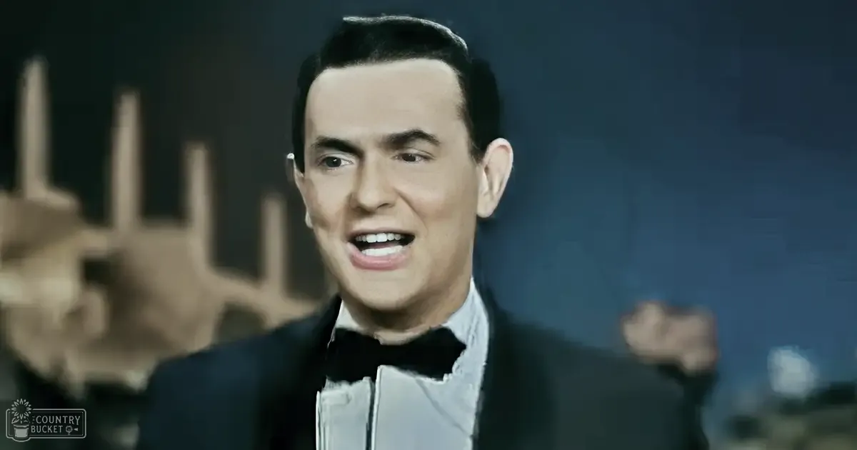 A young adult male wearing a formal tuxedo with a bow tie stands on stage in a dimly lit environment, likely during an event.
