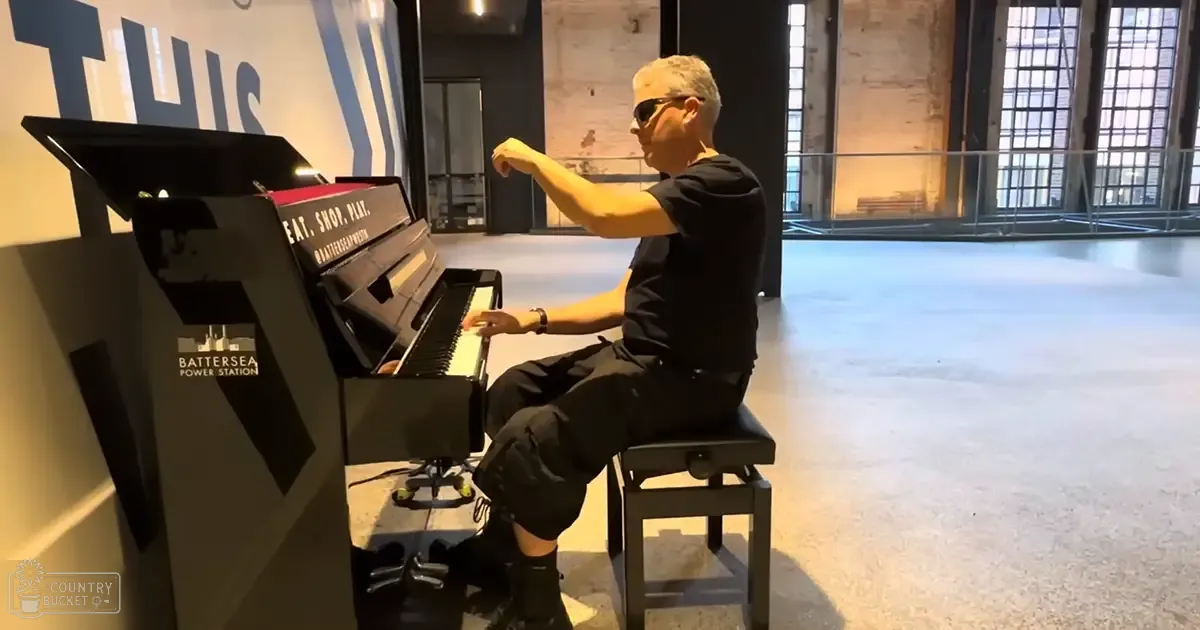 A middle-aged adult male plays a piano in Battersea Power Station, surrounded by large windows and an industrial interior with exposed brick walls.