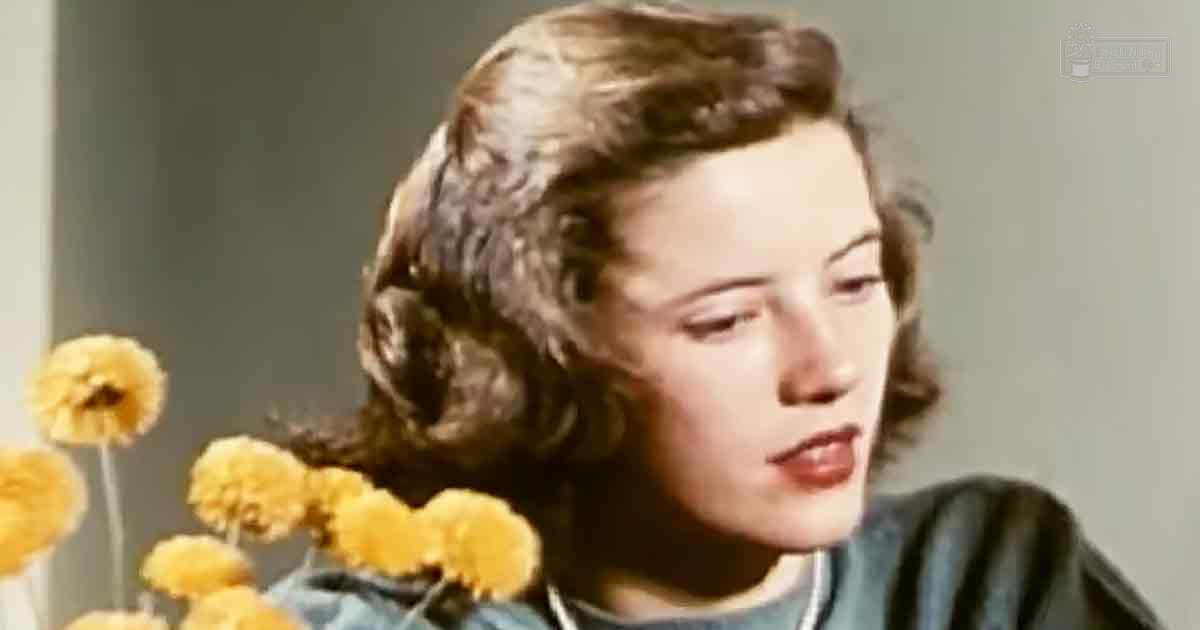 A young adult female wearing a dark green, mid-20th century-style garment indoors with muted background and soft lighting.