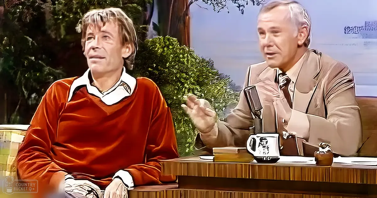 Two individuals seated at a desk in an indoor talk show setting with greenery and bright lighting.
