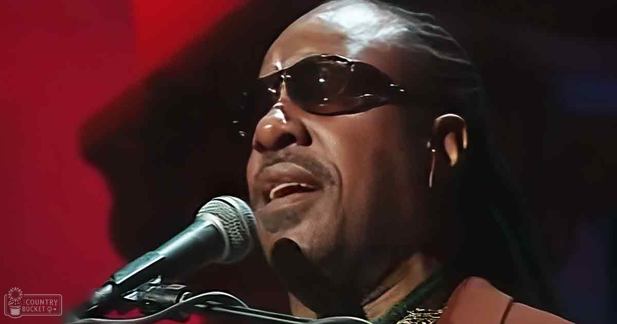 Adult male singer wearing sunglasses and a black garment with high collar performing on stage in front of a microphone at a concert or music event.