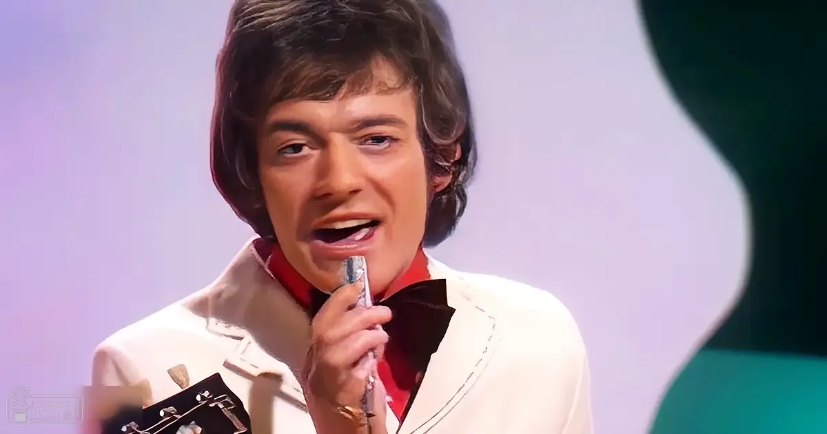 A young adult male in a white blazer with black lapels and red lining performs on stage under purple and white lighting, holding a microphone.