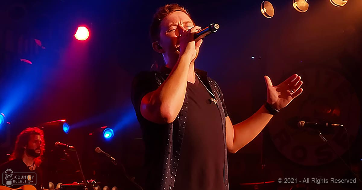 A young adult male singer in a dark, studded shirt and black vest performs at a concert with stage lights creating a dramatic red and blue ambiance.