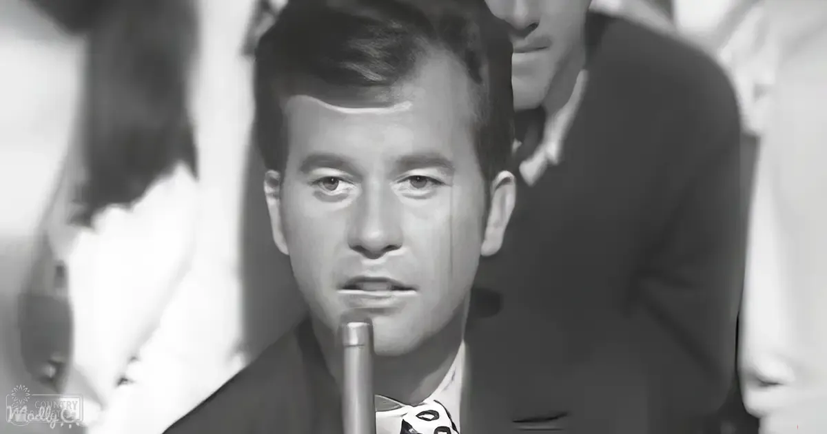 A young adult male in a dark suit with a patterned tie stands at a microphone indoors, possibly during a mid-20th century performance or radio broadcast.