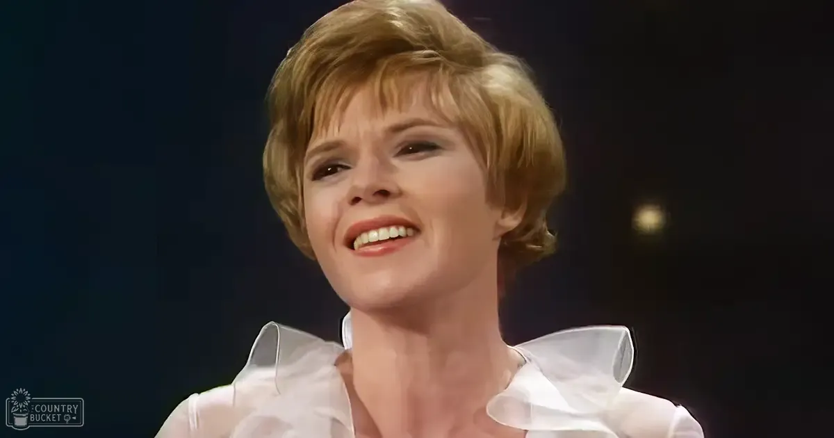 A middle-aged woman in an indoor setting wearing a white blouse with ruffled details around the collar and shoulders, possibly from mid-20th century style.