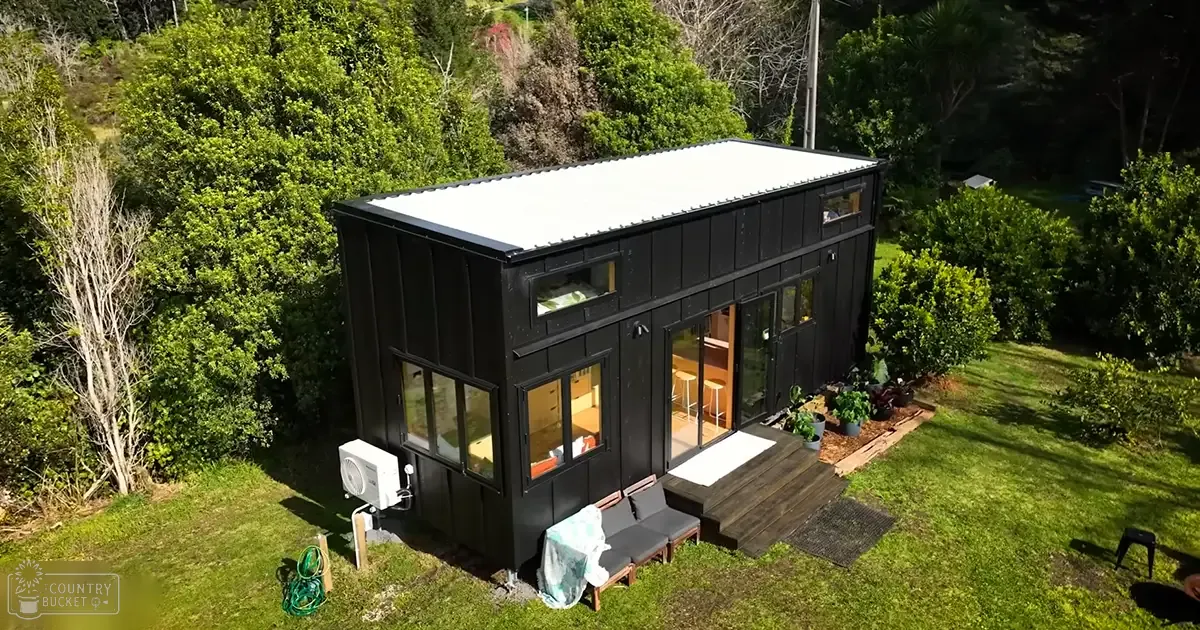 A modern black tiny house with white trim, situated in a lush green area surrounded by trees. The scene includes a wooden deck leading to the entrance and some furniture inside.
