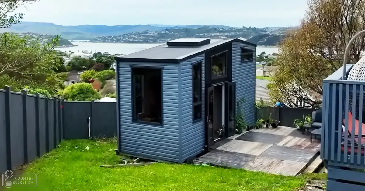 A suburban backyard scene with a modern tiny house, wooden deck, potted plant, grassy lawn, privacy fence, view of water and hills, spring or summer season, daytime light