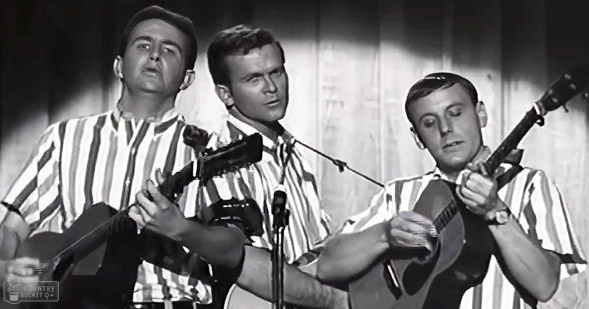 A musical performance scene with young adult males holding guitars and microphones in a simple indoor setting, possibly a club or small theater.