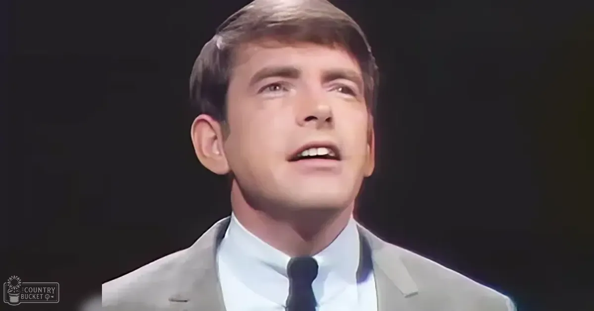 Young adult male in light gray suit jacket with white shirt, mid-20th century style, against dark background suggesting stage setting.