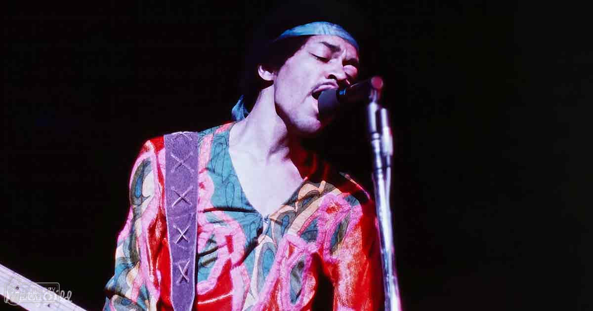Adult male singer wearing a vibrant, patterned shirt with reds, blues, and greens, performing on stage under dramatic lighting at what appears to be a concert.