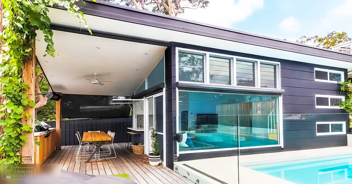 A suburban home with a modern design with a patio area, outdoor furniture, potted plants, ceiling fans, large sliding doors leading to a pool, and dark siding on the house.
