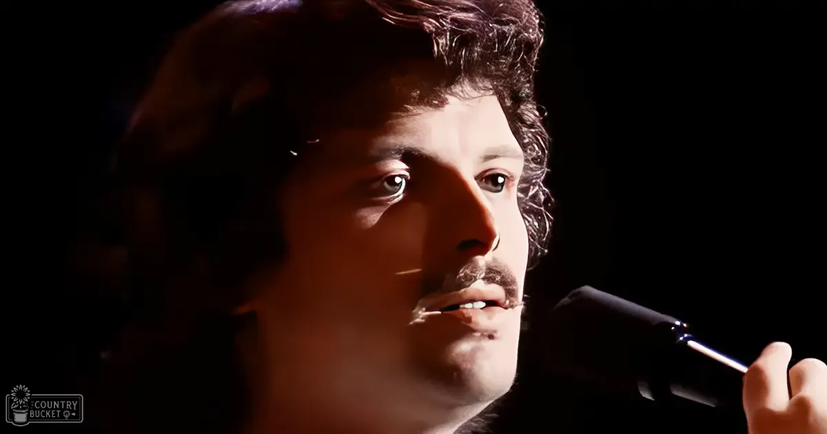A young adult male singer wearing a dark, textured knit garment from the late 1980s sings into a microphone on stage under focused lighting.