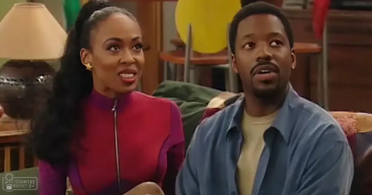 Adult female wearing a red zip-up top with gold hoop earrings, seated indoors; adult male in light blue button-up shirt over cream turtleneck sweater, sitting nearby. Living room setting with beige lamp shade and green cushioned chair.