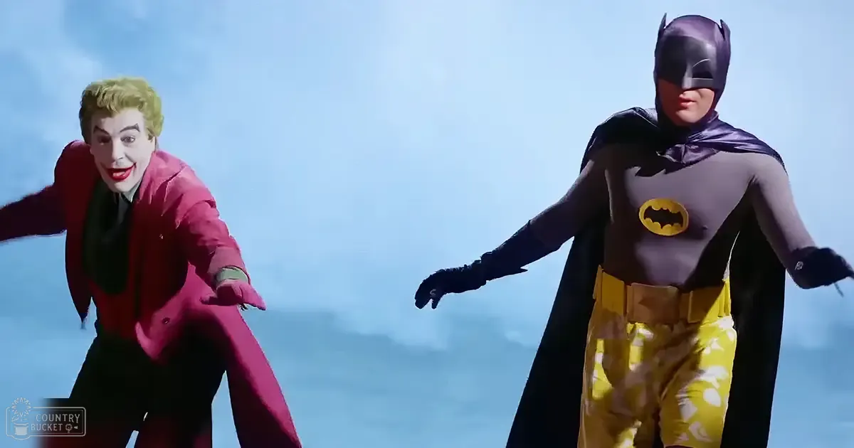Two adults in classic Batman costumes stand against a clear blue sky, possibly on a rooftop or elevated platform during the daytime.