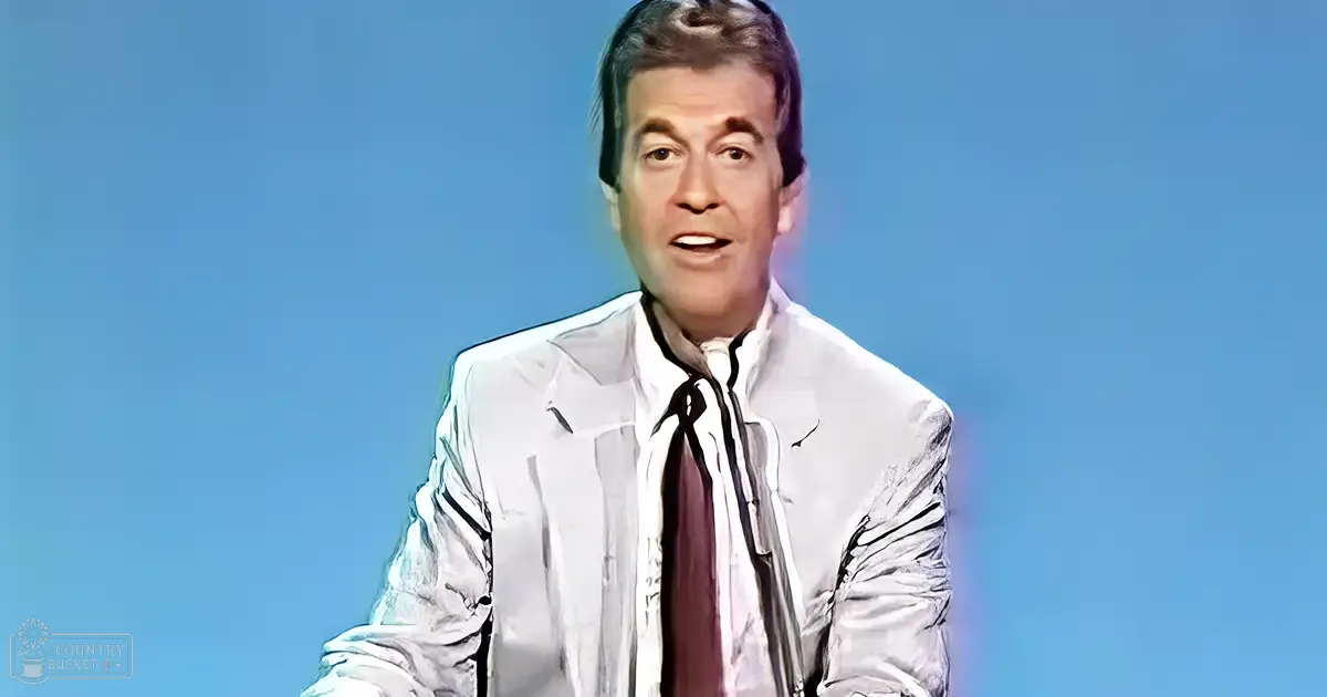 A middle-aged adult male in a formal white suit jacket against a plain blue indoor background.