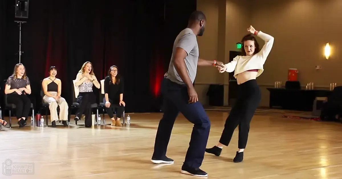 Indoor dance rehearsal with young adults in casual attire, audience of women in various outfits, stage lighting suggesting evening or night setting.