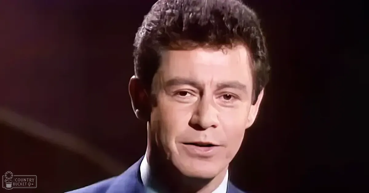 A middle-aged adult male wearing a dark blue suit jacket and white collared shirt in a dimly lit indoor setting, possibly on stage.