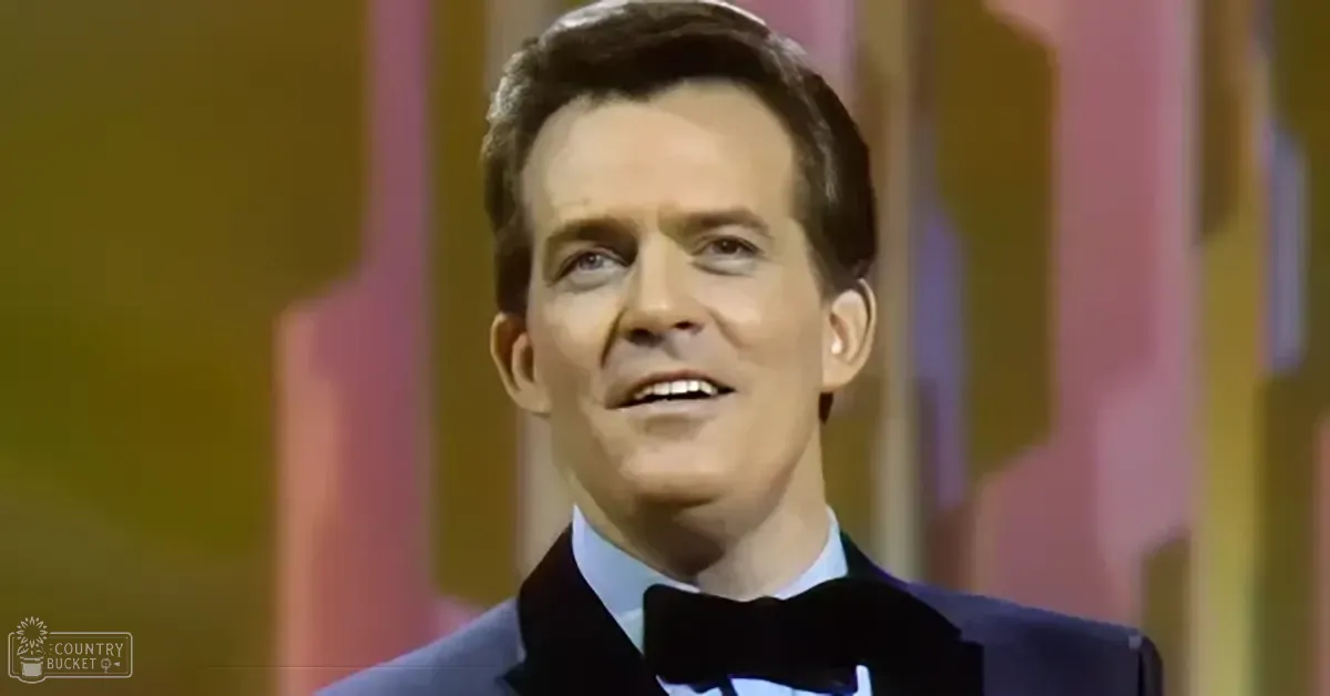A middle-aged man in a formal suit jacket with a bow tie stands against a colorful abstract backdrop indoors.