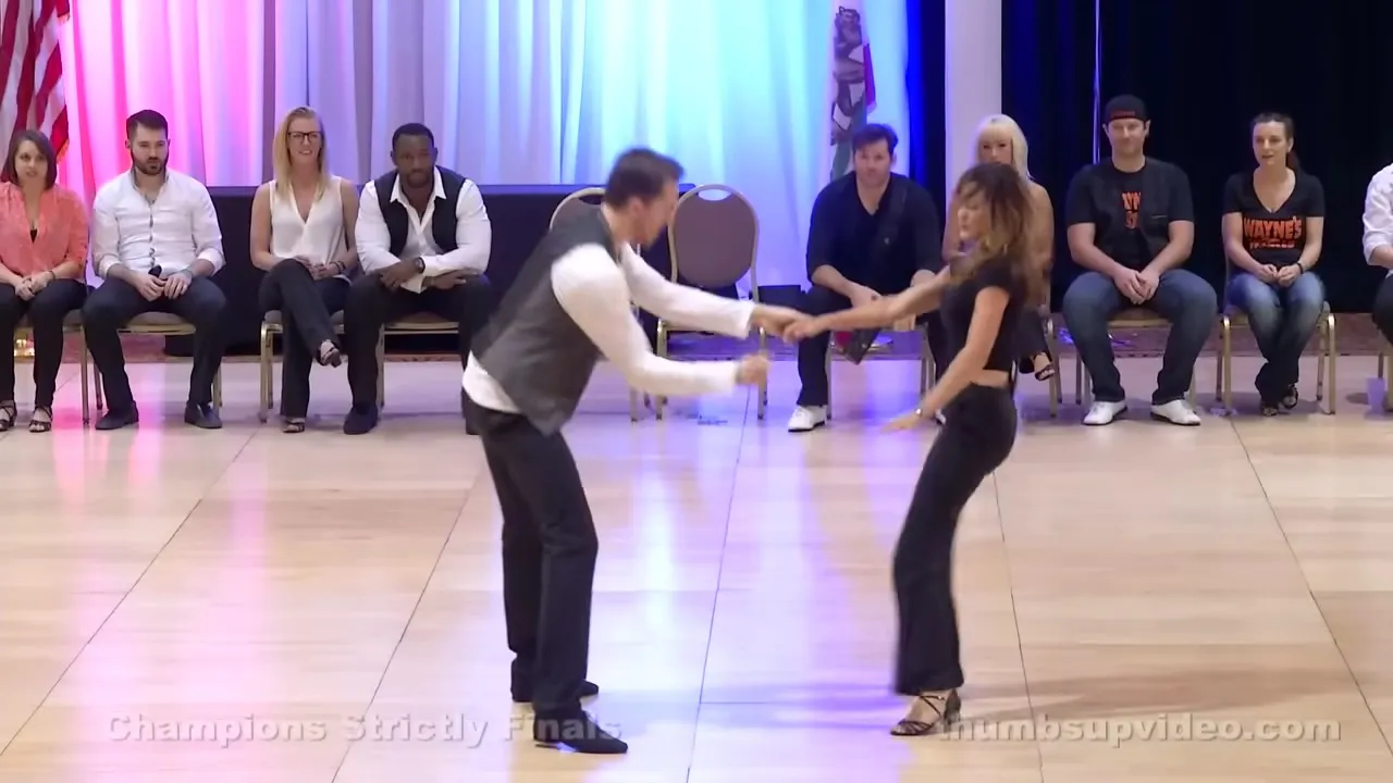 A lively dance performance at "Champions Strictly Finals" in an indoor ballroom, with young adult dancers and spectators in casual attire.