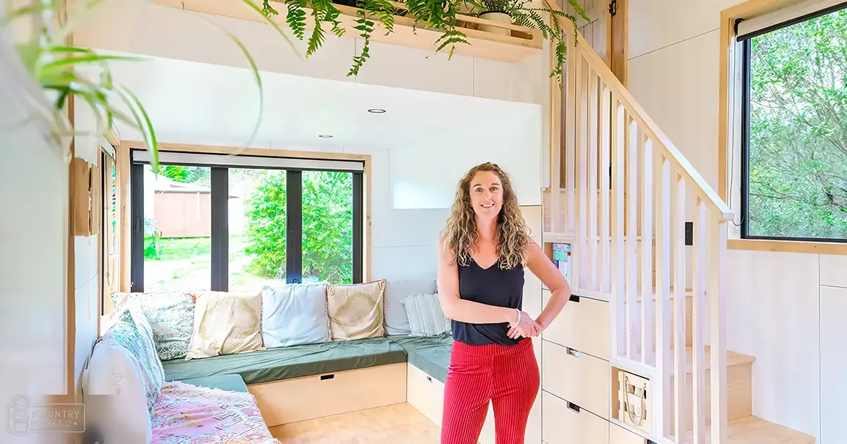 A young adult female in a modern, casual outfit of a black sleeveless top with a v-neckline and red corduroy pants, surrounded by indoor furniture such as a cushioned bench with green and patterned pillows, white railings leading to an upper level, wooden storage units beneath the stairs, and hanging plants adding natural light. The setting appears to be indoors within a tiny house during daytime in spring or summer.