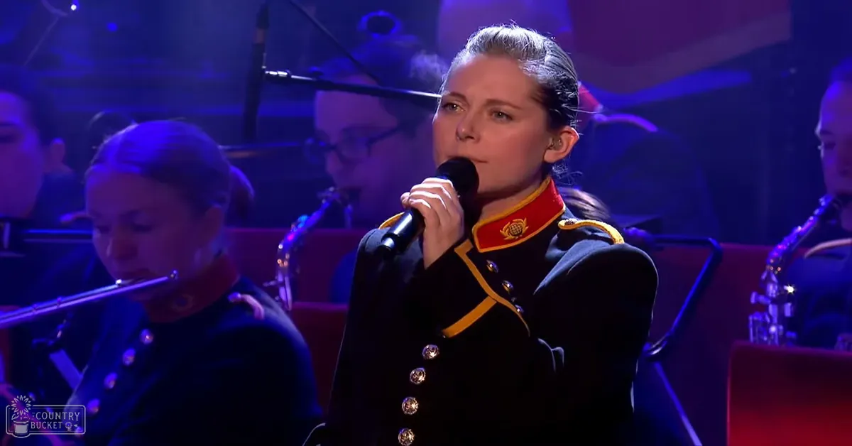 A young adult female in a dark blue military-style jacket sings into a microphone on stage with other musicians playing instruments in an indoor concert hall setting.