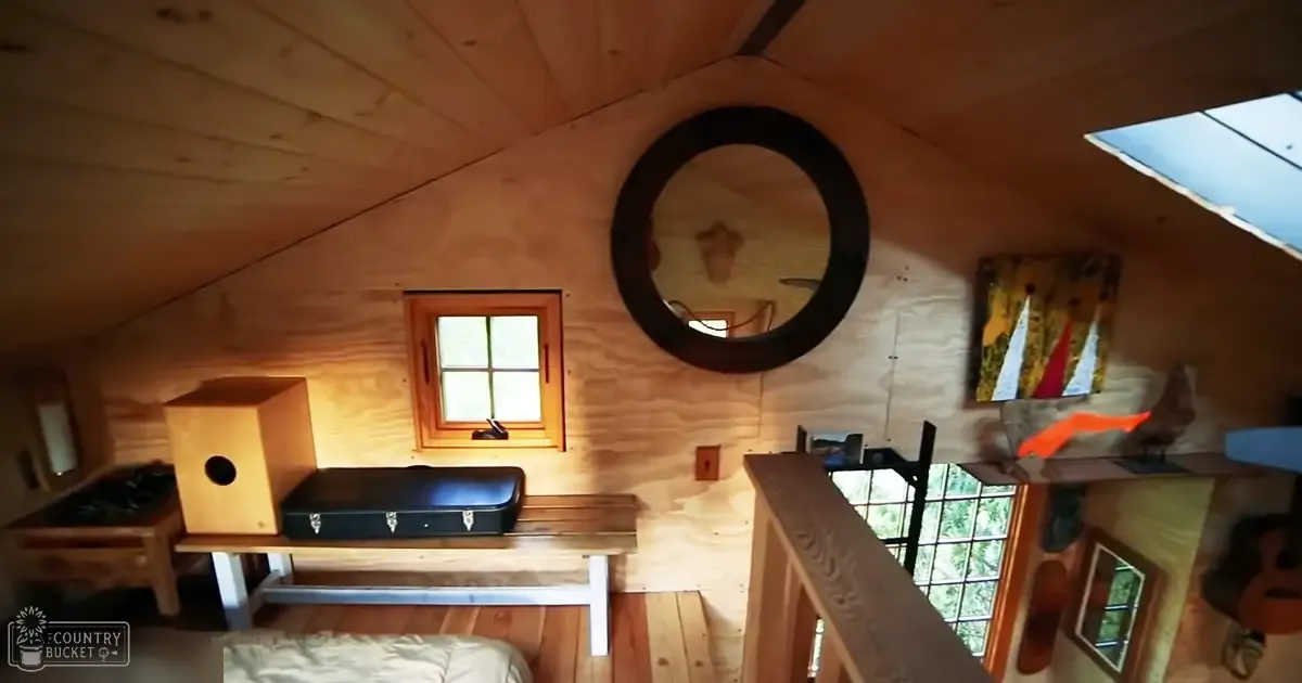 Cozy attic or loft interior space, wooden bench with black suitcase, small wooden box with circular cutout, round mirror hanging above, painting leaning against wall to right, warm tones of beige, brown, and white walls and ceiling, natural light from window suggests daytime setting