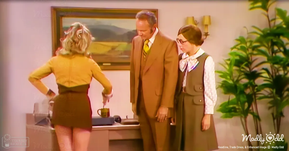 An adult female in a yellow sleeveless top and dark skirt, standing in an office environment with other individuals. The scene includes furniture like a desk and wall-mounted lamps.